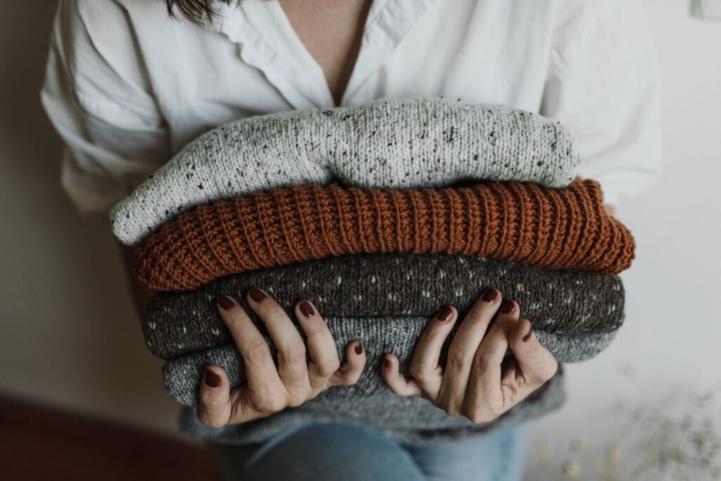 A person holding a stack of folded knitted sweaters, the kind of garment a delicate cycle exists for