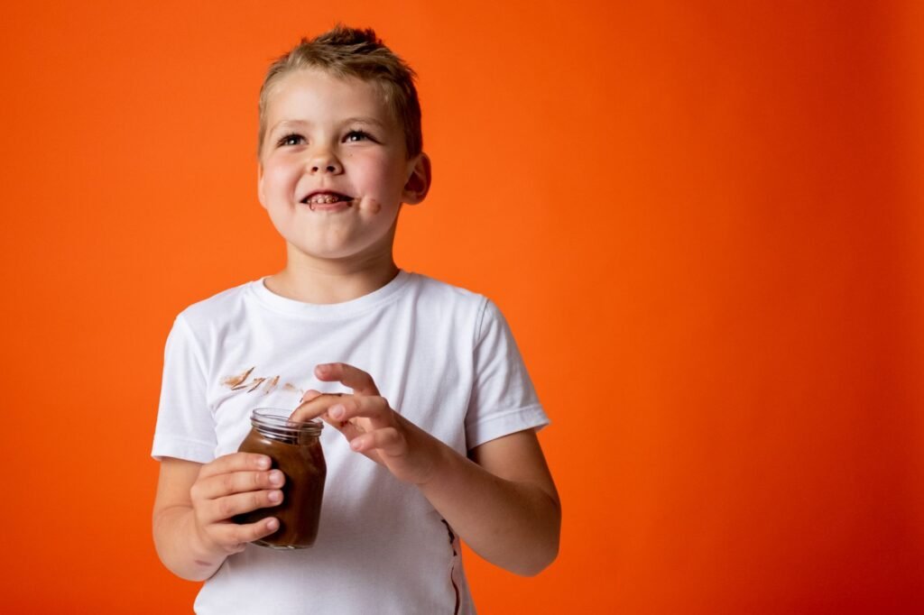 Child in a white t-shirt holding a jar of chocolate spread with chocolate smeared down the front of the shirt
