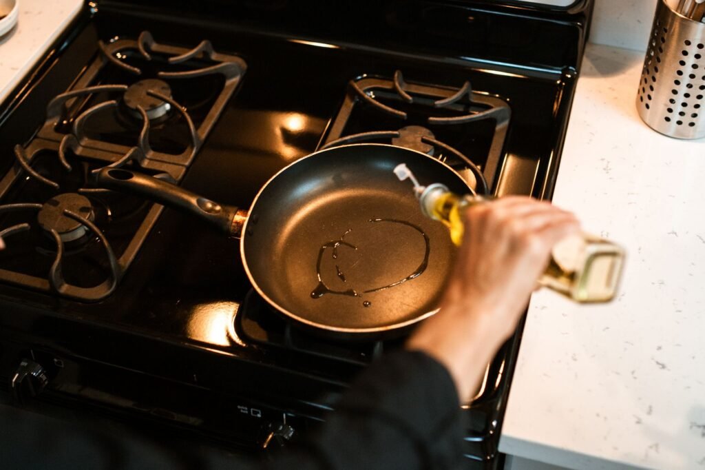 Cook pouring olive oil into a hot frying pan on a stove, the moment most cooking oil stains on clothes begin