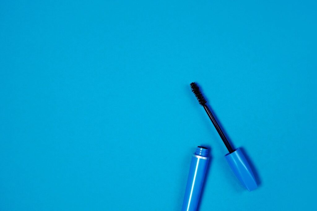 Close-up of an open mascara tube and wand against a blue background, the applicator that transfers most mascara stains to clothing