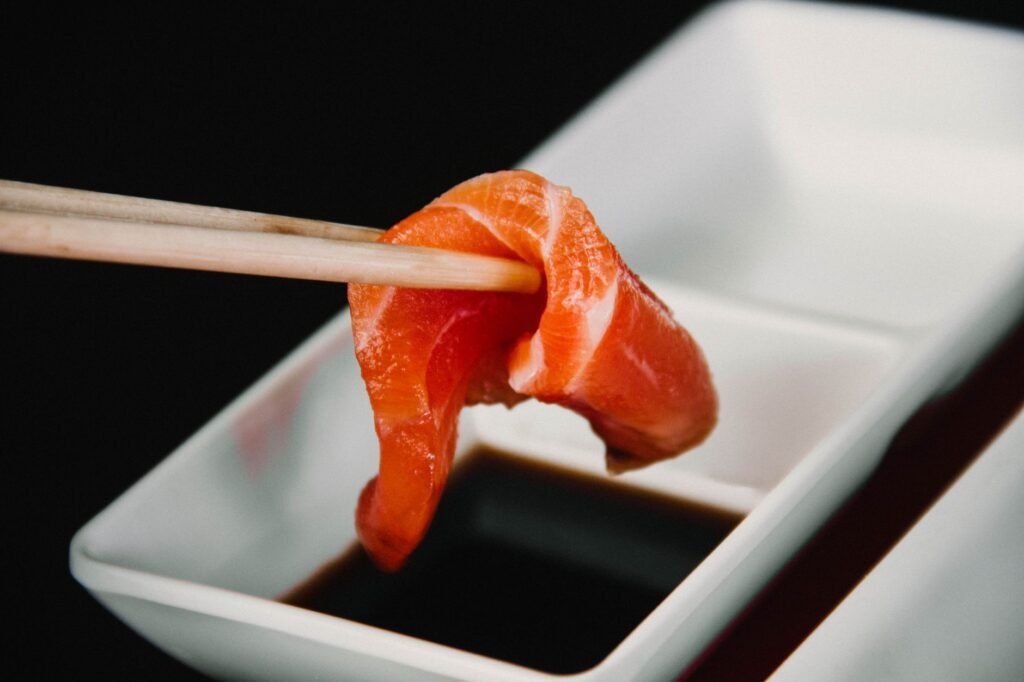 Piece of salmon sashimi held in chopsticks being dipped into a dish of dark soy sauce