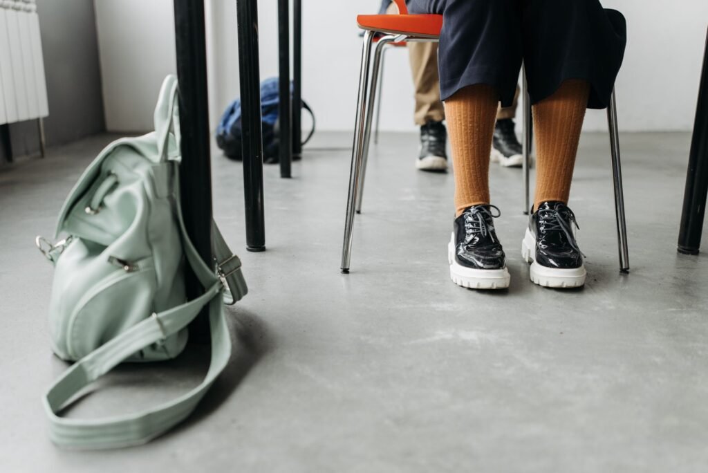 A pale green backpack on the floor beside a school desk, straps and buckles loose
