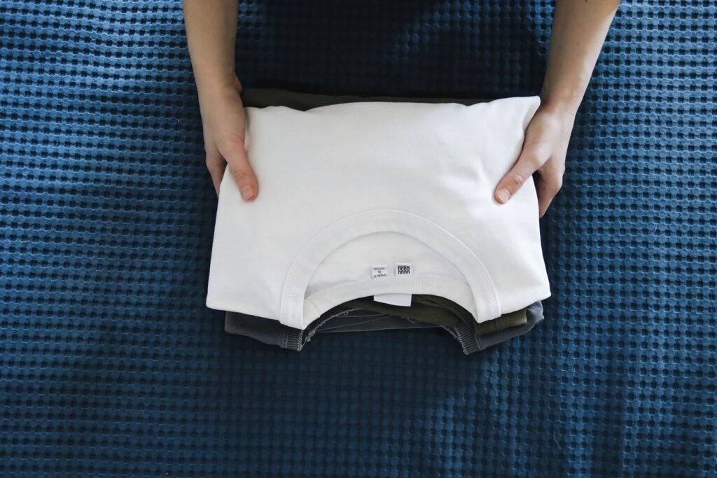 Hands placing a folded white shirt on top of a stack of darker clothes on a blue bedspread