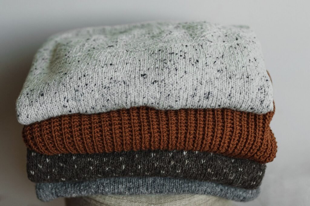 A stack of folded knitted jumpers in grey, rust and cream, the kind of fabric that both sheds lint and collects it