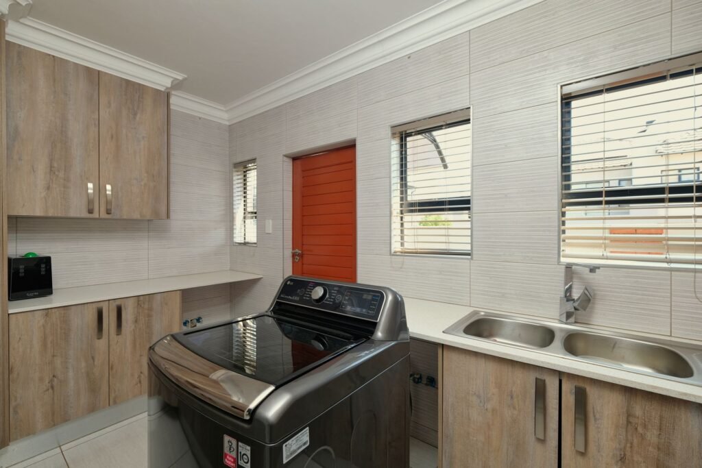 A laundry room with a top-load washing machine standing next to a double stainless steel sink, the two fixtures that share a drain