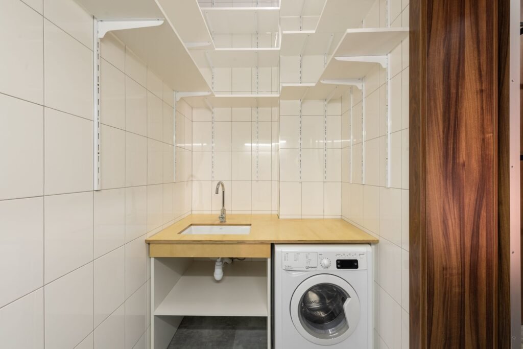 A narrow tiled utility room with a front-load washer built in beneath a worktop and sink