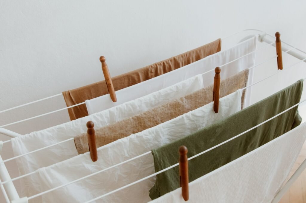 An indoor concertina drying rack with white, linen, rust and olive items draped over its rails and wooden clothespins clipped along the bars against a plain wall