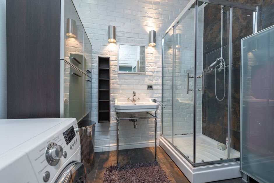 A white front-load washing machine in the foreground of a white-brick bathroom with a glass corner shower and a basin on chrome legs