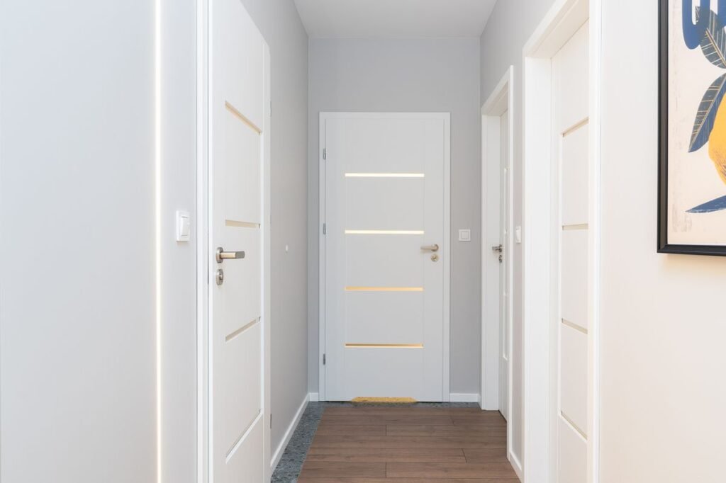 A narrow white apartment hallway with several closed internal doors and a wooden floor, the route any apartment size washer has to travel