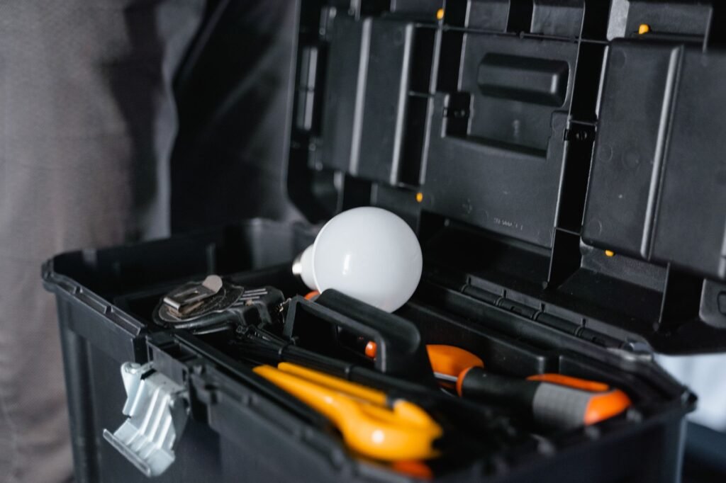 Open black plastic toolbox with a lift-out tray holding pliers, an orange-handled screwdriver, an adjustable wrench and a white light bulb