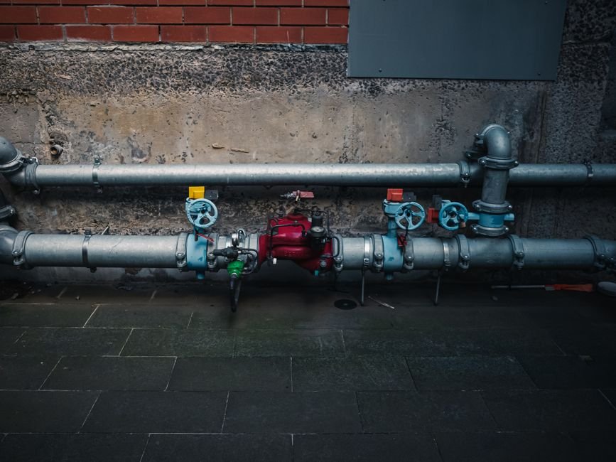 Steel water pipes on a wall fitted with blue handwheel shutoff valves and a red backflow preventer, the kind of main line an automatic water shutoff valve installs on