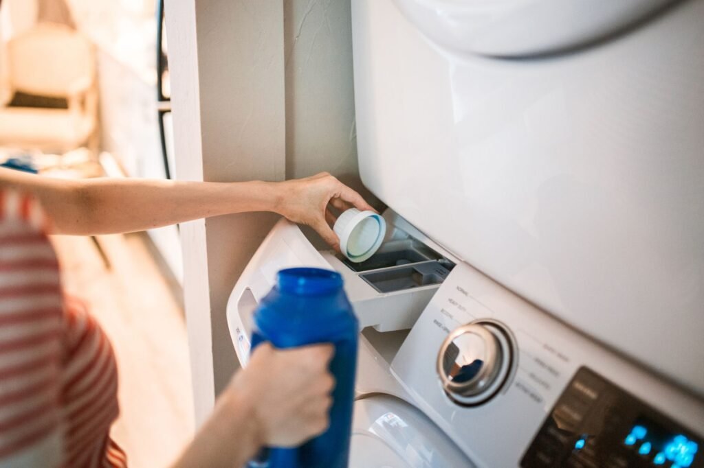A hand tipping a capful of powder into the open dispenser drawer of a front-loading washer with a dryer stacked above it, a blue detergent bottle in the other hand