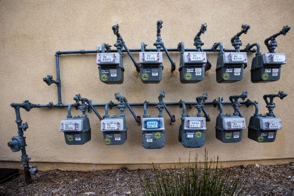 Eleven domestic gas meters in two rows on a cream stucco wall, each fed by grey steel pipework with its own shutoff cock and marked with a yellow call-before-you-dig sticker