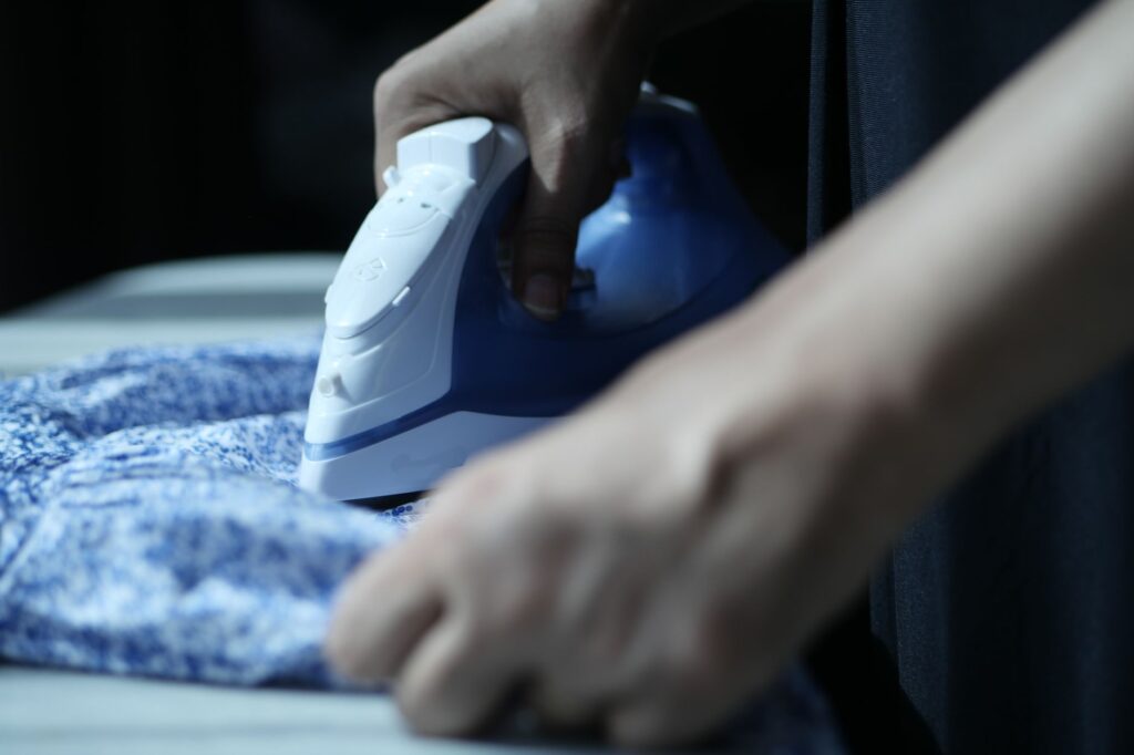 Close-up of a blue and white steam iron being pushed across patterned blue fabric with a hand steadying the cloth ahead of the soleplate