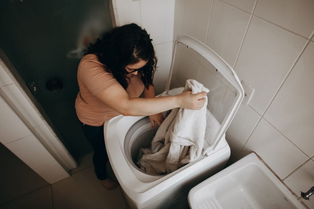 Seen from above, a woman lowering towels into an open top-loading washing machine in a white-tiled bathroom, the lid raised and a utility sink beside it