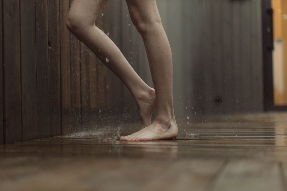 Bare feet standing on a wet dark wooden floor with water splashing around them, the indoor flooding a water leak detector exists to catch early