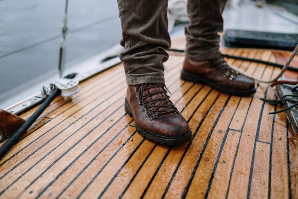 Two soaked brown leather hiking boots with the water beaded on them, standing on a wet varnished teak deck beside coiled rope