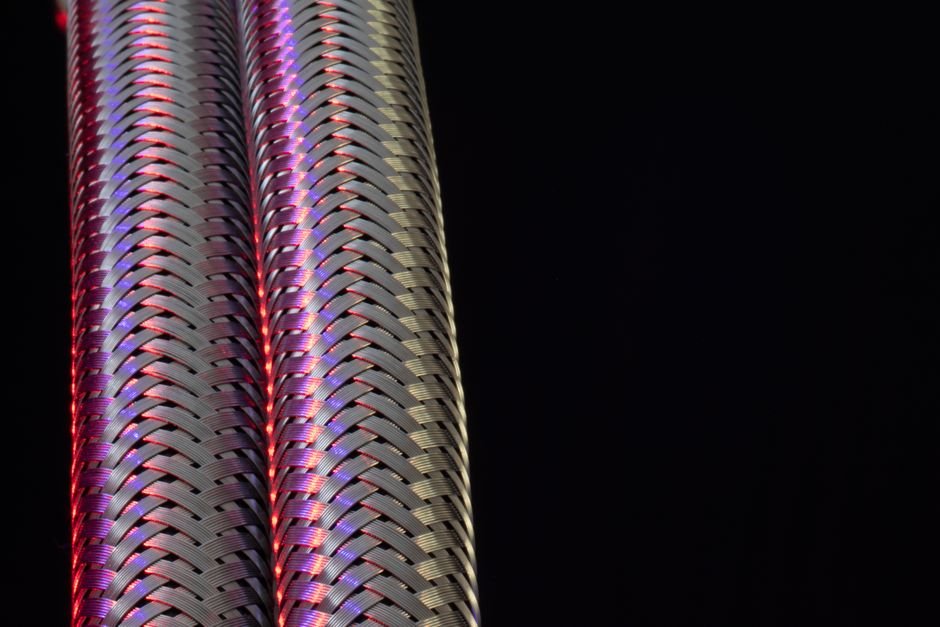Close-up of woven stainless steel hose braid lit in red and violet against black, the outer layer that gives braided washing machine hoses their burst strength