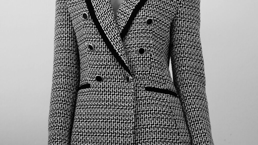 A tailored black and white houndstooth tweed jacket on a dress form, double-breasted with black velvet trim on the lapels and pockets, shot against a plain grey backdrop