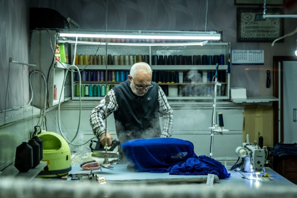 A tailor presses blue fabric on a workshop table with steam rising from the cloth, a green steam generator canister beside him and racks of thread cones behind