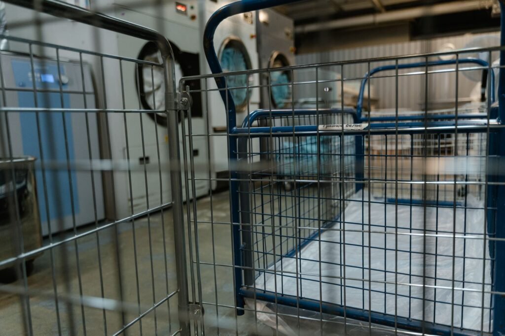Wheeled wire laundry trolleys lined up on a wet concrete floor in a commercial laundry, with large industrial front-loading machines and a control panel behind them