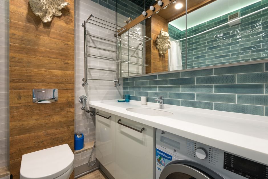 A white front-load machine slotted under a solid worktop beside a bathroom vanity, with teal metro tiles behind and a chrome ladder rail on the wall, a typical compact dryer installation
