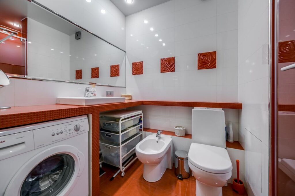 A white front-load washing machine built in beneath an orange tiled worktop next to a basin in a narrow white and orange bathroom