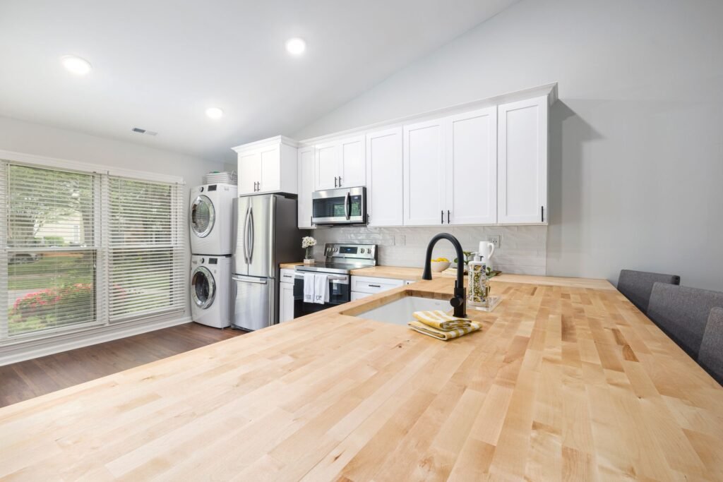 A long bare maple butcher-block worktop with an undermount sink and black tap in the foreground, white wall cabinets above and a stacked washer and dryer against the far wall