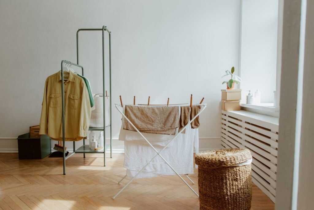 A folding clothes airer holding damp sheets and a towel in a bare room with a herringbone wood floor, a clothes rail behind it and a wicker laundry basket beside a radiator