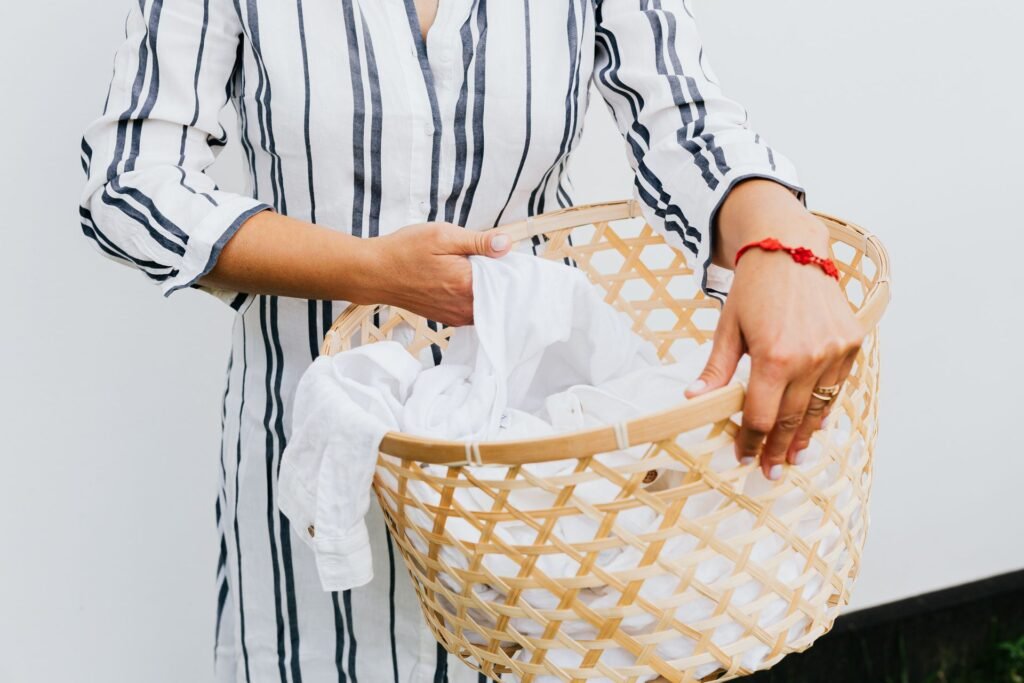 A woman in a navy-striped shirt dress carrying a woven bamboo basket of white laundry against a plain white wall
