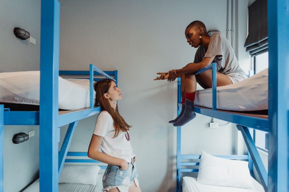 Two students talking in a residence hall room with blue steel bunk beds, one sitting on the top bunk in socks, the kind of shared space a dorm washing machine has to fit into