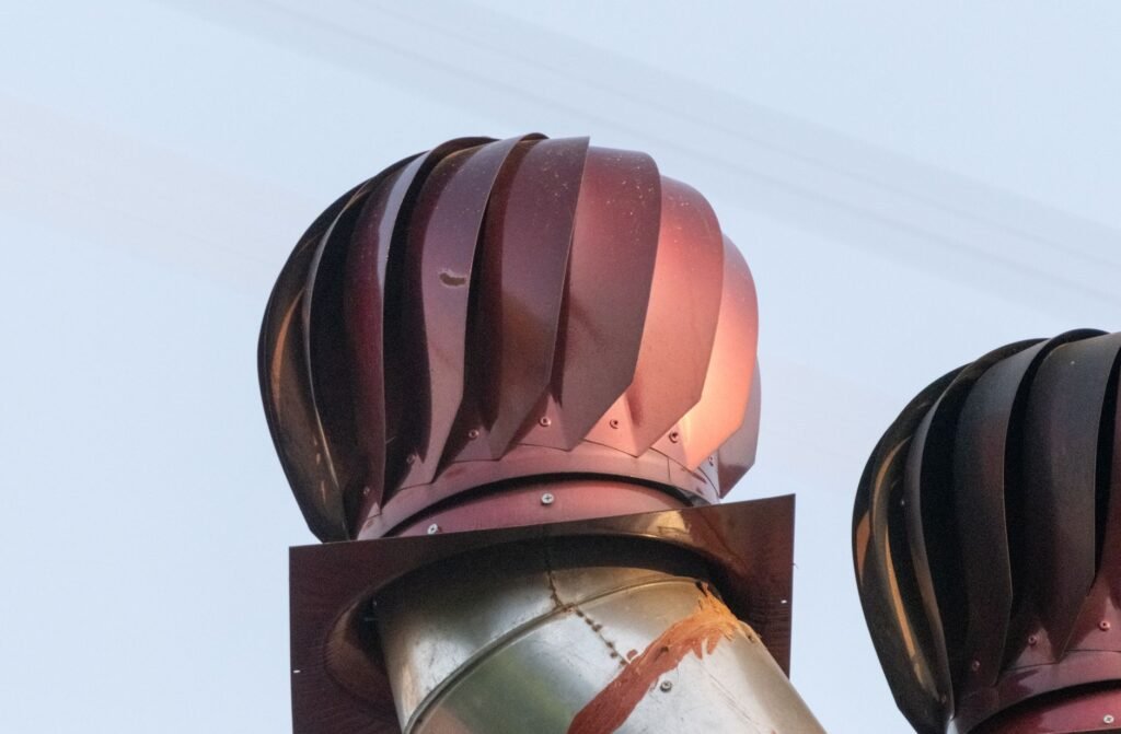 A roof turbine ventilator with curved overlapping metal vanes mounted on top of a galvanised steel duct against a pale sky, a second one beside it