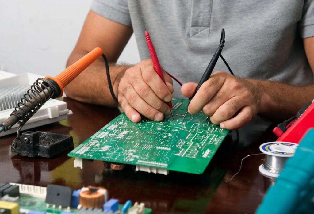 A technician holding two multimeter probes against a large green printed circuit board on a bench, with a soldering iron, solder spool and a second board beside it