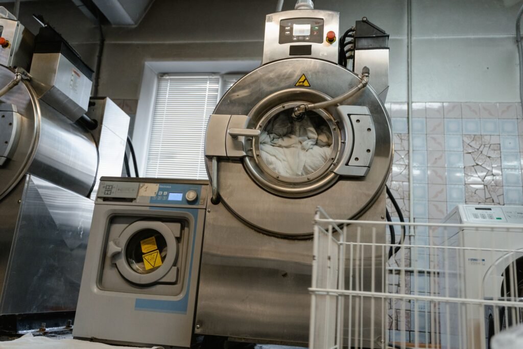 A large industrial laundry machine with its heavy round door swung wide open on its hinge, drum full of white linen, beside a smaller front loader with its door ajar