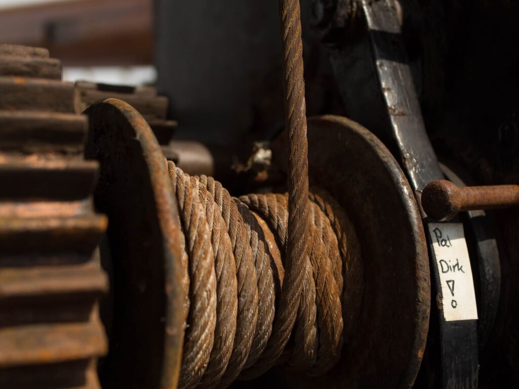A heavily rusted steel winch drum wound with wire rope, turning on its shaft beside a worn cast gear wheel in dim workshop light