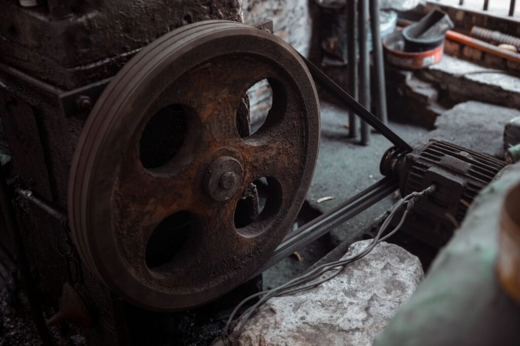 A large cast-iron pulley wheel on an old workshop machine, driven by a narrow belt running down to a small electric motor bolted to the floor