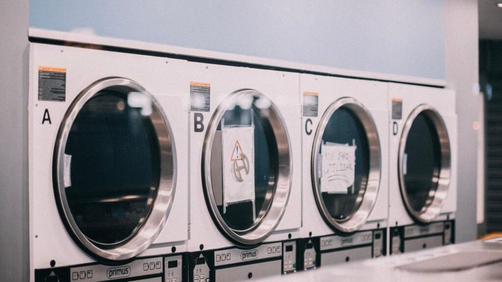 Four white commercial tumble dryers lettered A to D in a laundrette, two of them with handwritten out-of-order notices taped inside the glass doors