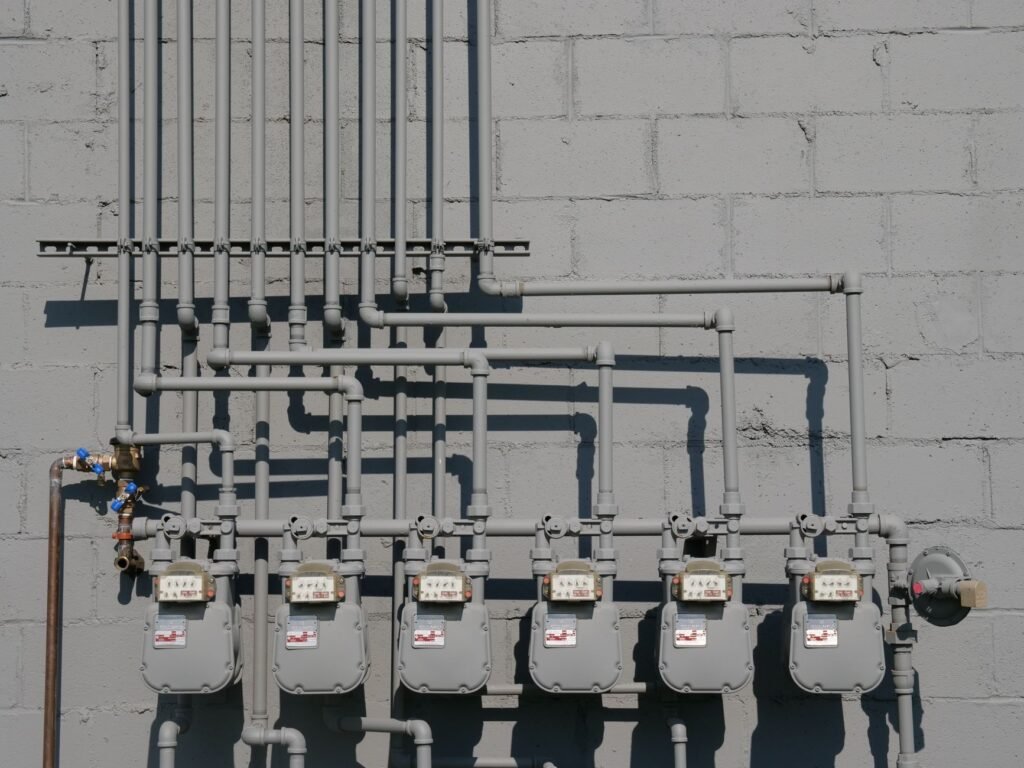 Six grey domestic gas meters mounted in a row on a painted block wall, fed by steel pipework with shutoff valves rising to the ceiling