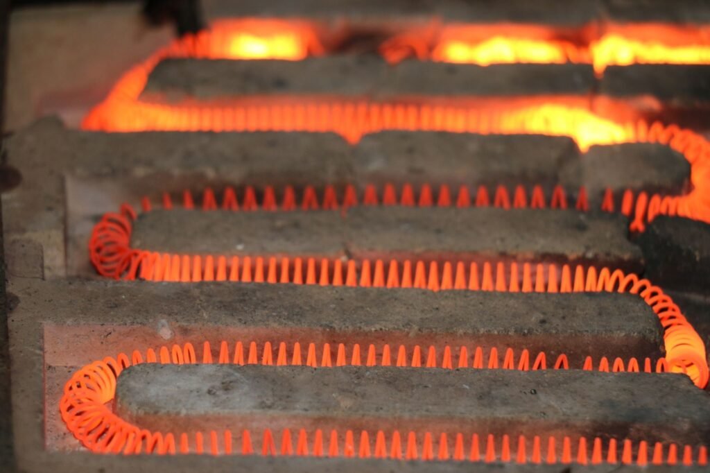 A coil of resistance wire glowing bright orange as it snakes across grey firebrick inside a heating chamber, with more glowing coils behind it