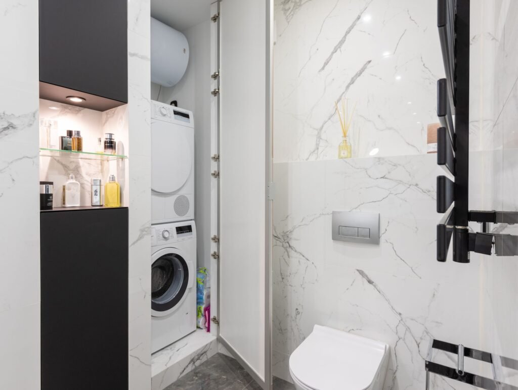 A condenser dryer stacked above a front-load washer inside a narrow cupboard in a marble bathroom, with a wall water heater above them