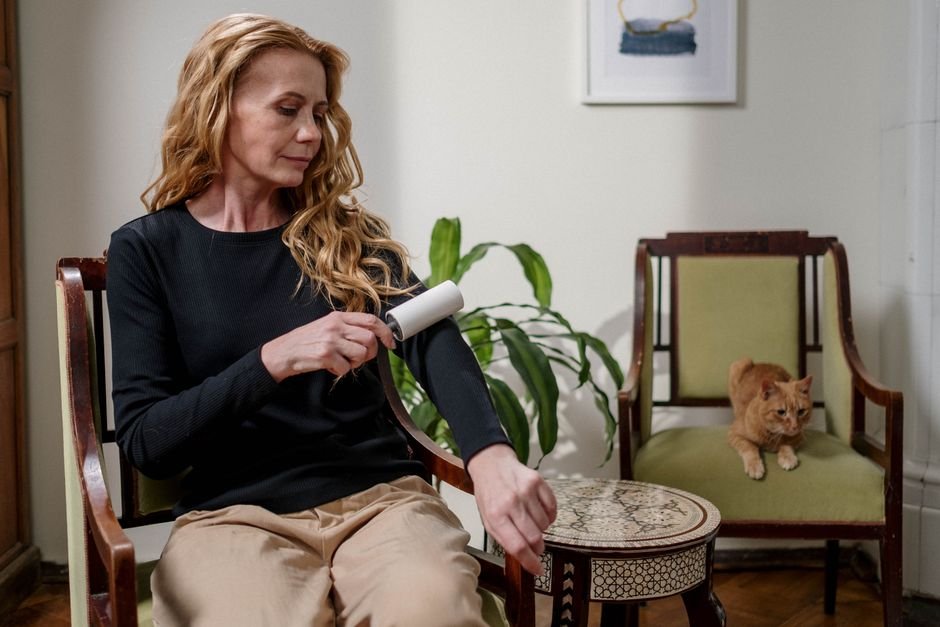 Woman in a black top running a lint roller down her sleeve in a sitting room, a ginger cat watching from the chair opposite