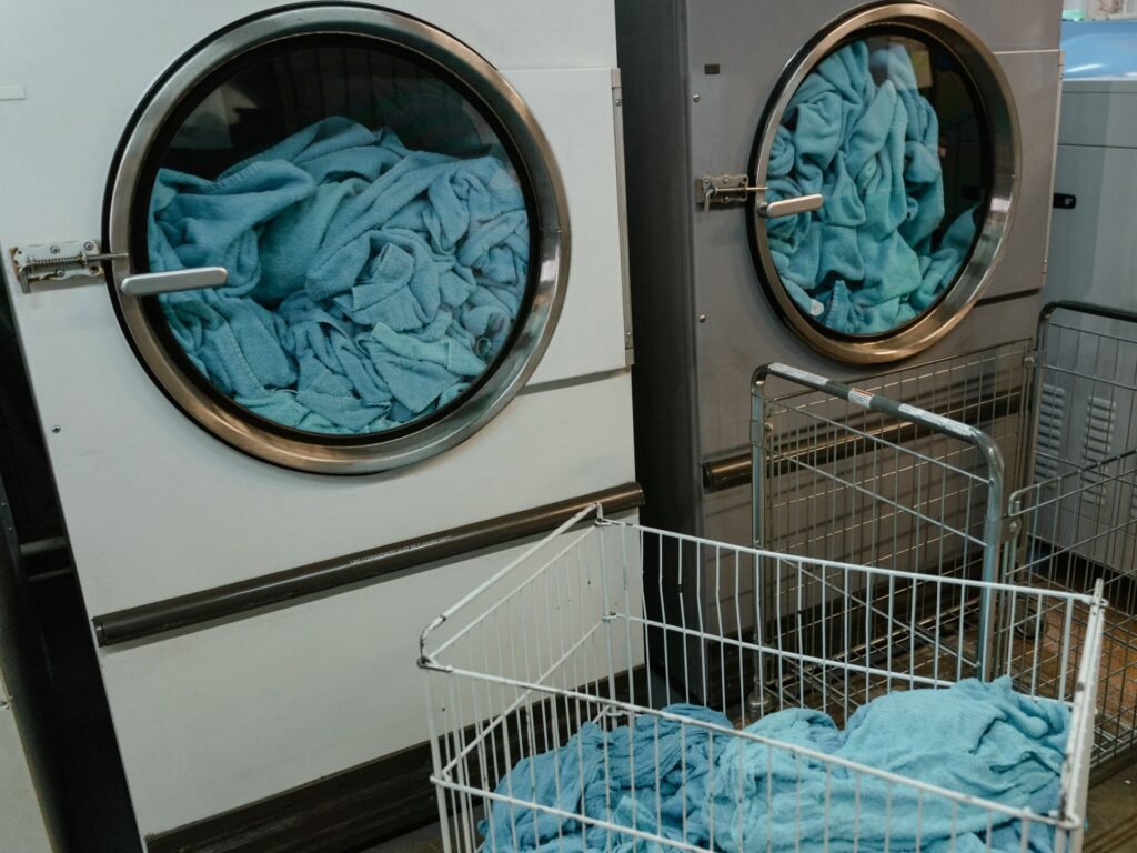Two commercial dryers with round glass doors packed tight with bunched-up pale blue towels, a wire laundry cart of more towels in front