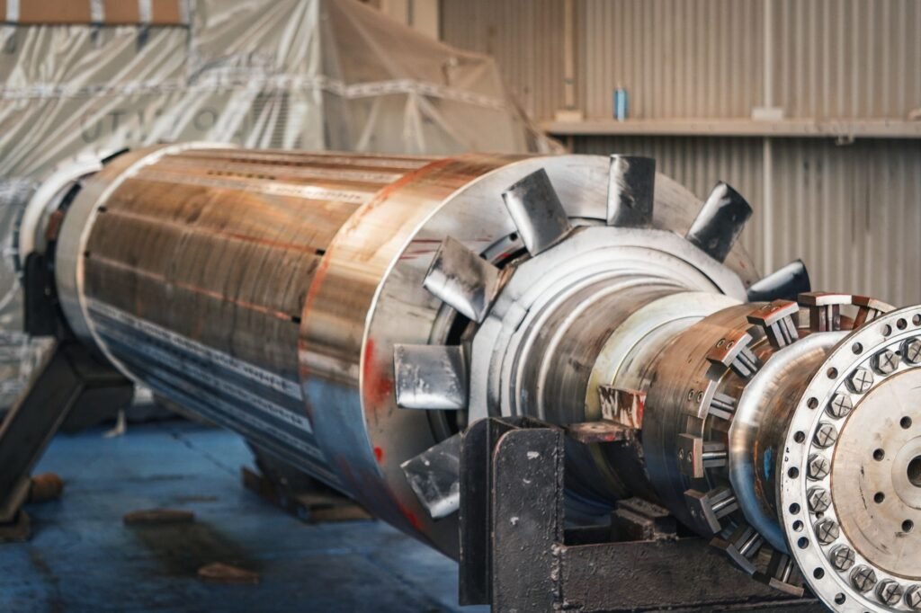 A large industrial electric motor rotor resting on steel stands in a workshop, its laminated core, cooling vanes, shaft and bolted end flange all exposed