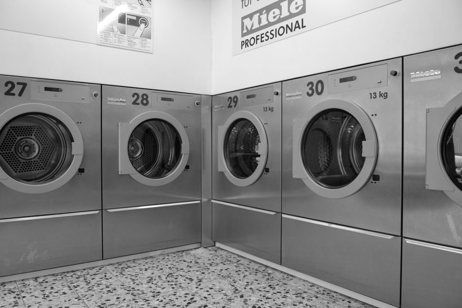 Black and white view of a row of numbered 13 kg commercial laundry machines with their doors ajar, showing the perforated drums inside, standing on a terrazzo floor