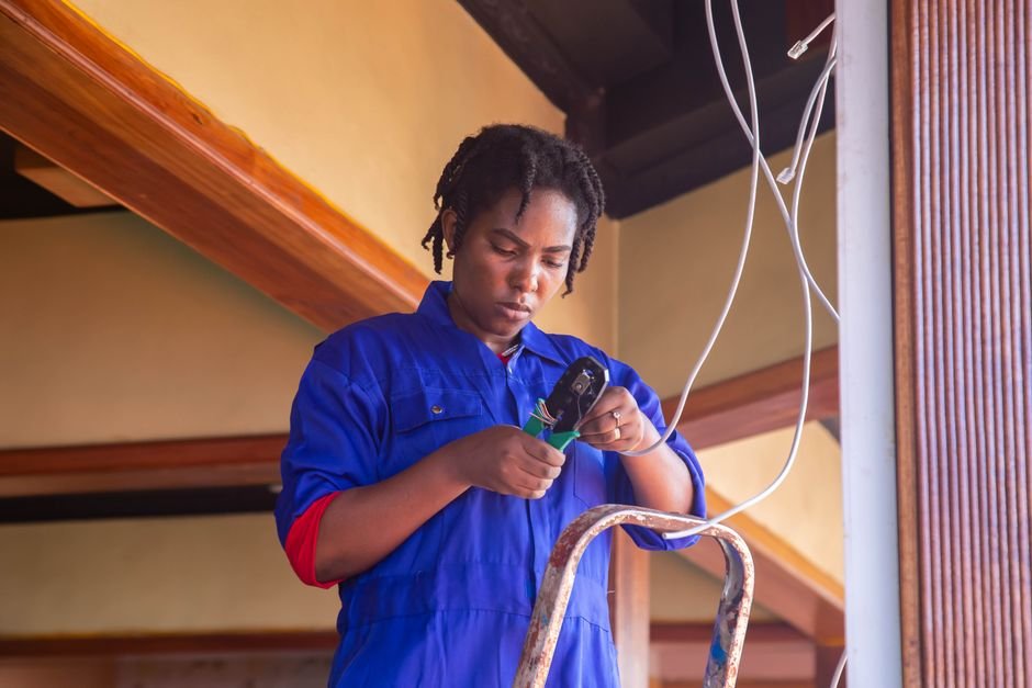 An electrician on a stepladder stripping the end of a multi-core cable with a wire stripper, with loose cable hanging from the ceiling behind her