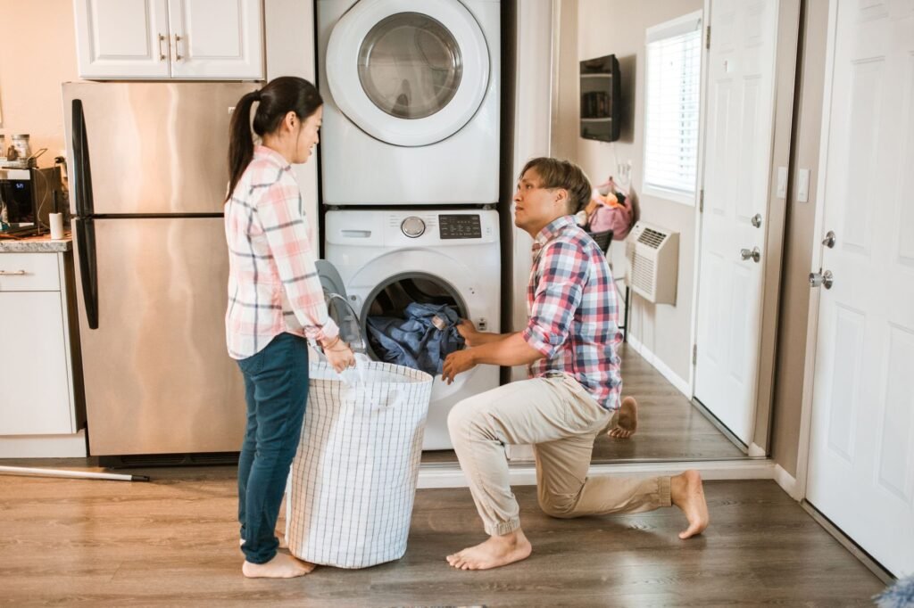 A couple at a stacked dryer and washer in a kitchen laundry nook, the man kneeling to lift jeans out of the open lower drum while the woman holds a mesh laundry hamper