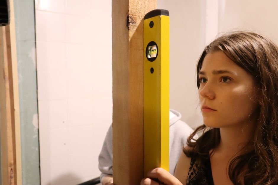 A woman holding a yellow spirit level flat against a timber upright indoors, watching the bubble in the central vial to check whether it is true