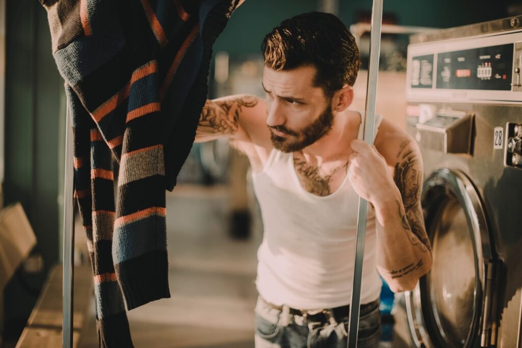 A man standing beside an open commercial dryer in a laundromat, examining a striped knitted sweater hanging on a rail in front of him with an unconvinced expression