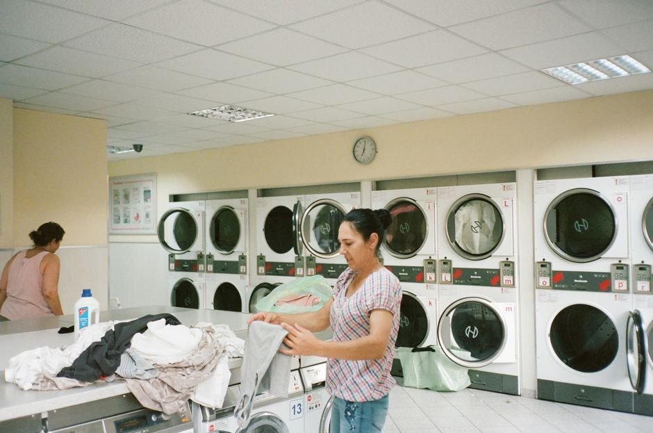 A laundrette with two walls of stacked tumble dryers, each with its own lettered selector and coin slot, a clock on the wall above them and a woman folding laundry in the middle of the room