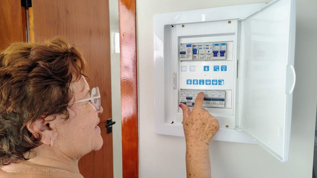 An older woman pressing a breaker switch inside an open domestic consumer unit mounted on a white wall, two rows of blue and white breakers visible behind the hinged cover